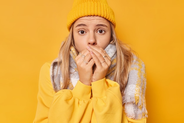 Bezpłatne zdjęcie młoda ładna kobieta czuje przeziębienie, próbuje się ogrzać, oddycha na rękach drży podczas mroźnej pogody, nosi czapkę i luźny sweter na żółtym tle. spaceruj w niskich temperaturach