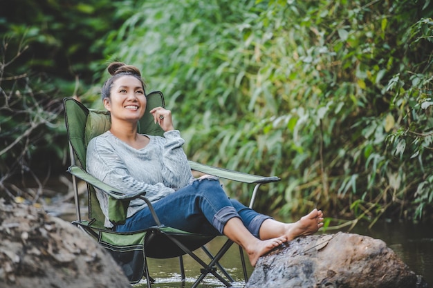Młoda ładna kobieta siedzi na krześle kempingowym w strumieniu dla relaksu, uśmiecha się w naturalnym lesie podczas podróży na kemping ze szczęściem miejsca kopiowania