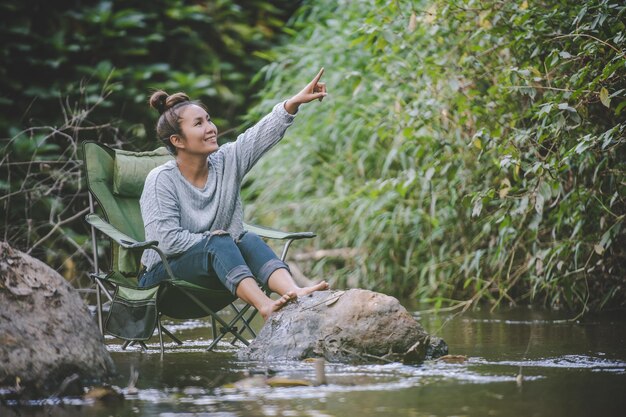 Młoda ładna kobieta siedzi na krześle kempingowym w strumieniu dla relaksu, uśmiecha się w naturalnym lesie podczas podróży na kemping ze szczęściem miejsca kopiowania