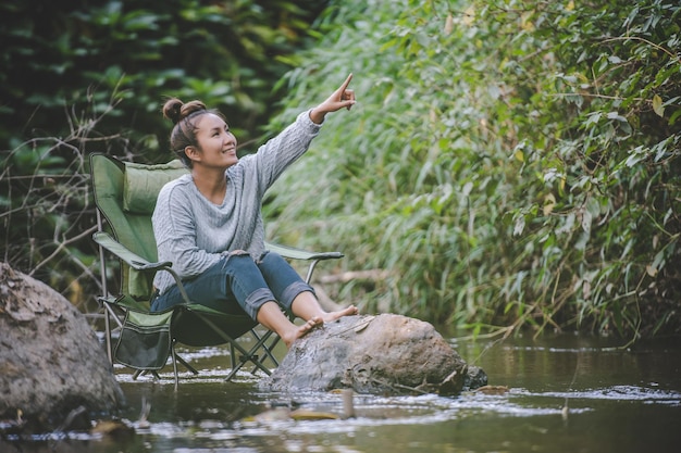 Młoda ładna kobieta siedzi na krześle kempingowym w strumieniu dla relaksu, uśmiecha się w naturalnym lesie podczas podróży na kemping ze szczęściem miejsca kopiowania