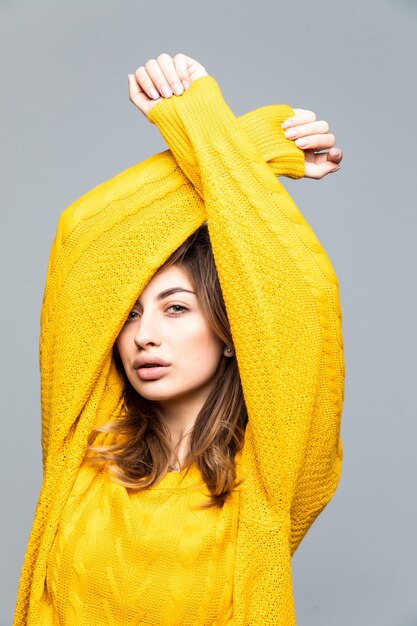 Młoda ładna kobieta w żółtym swetrze z uniesionymi rękoma na szarym tle ściany