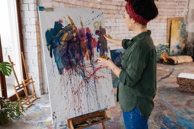 Młoda ładna kobieta z ciemnymi kręconymi włosami marzycielsko rysuje obraz na płótnie jasnymi farbami, spędzając czas w dużym przytulnym warsztacie
