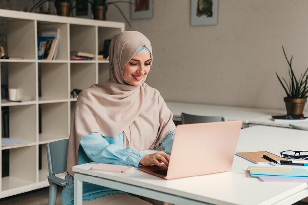 Młoda, ładna, nowoczesna muzułmanka w hidżabie, pracująca na laptopie w pokoju biurowym, edukacja online