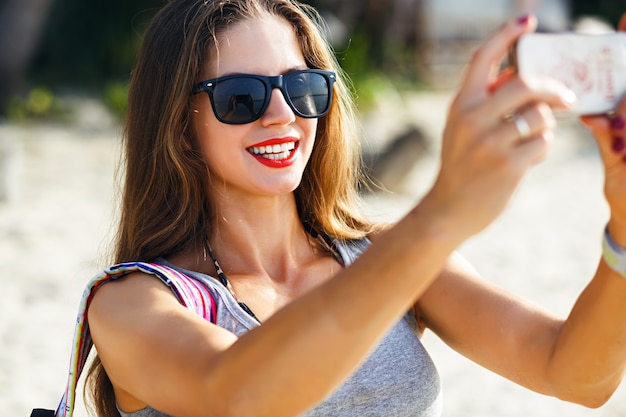 Młoda ładna podróżniczka robi zdjęcia na słonecznej plaży, podróżuje samotnie z plecakiem po gorącym tropikalnym kraju, swobodny strój, fitness body, przygodowy nastrój.