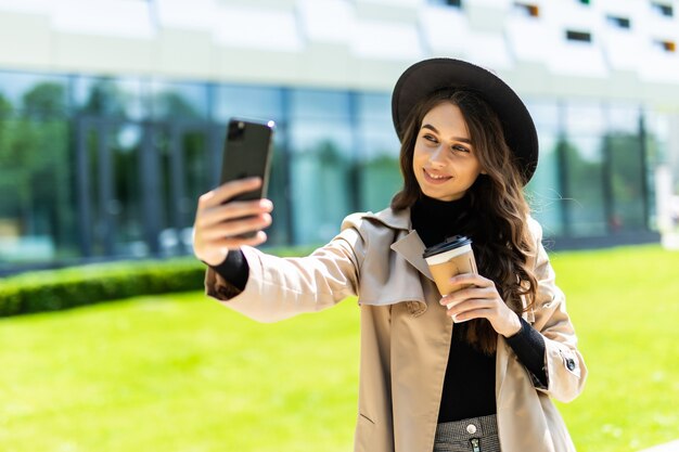 Młoda ładna studentka nosić płaszcz i kapelusz, trzymając kawę, aby wziąć selfie na telefon na ulicy