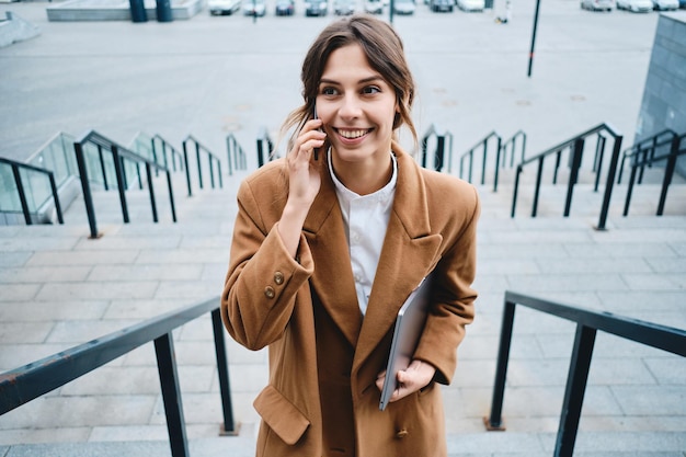 Młoda ładna uśmiechnięta bizneswoman w płaszczu z laptopem szczęśliwie rozmawiająca przez telefon komórkowy na schodach na zewnątrz