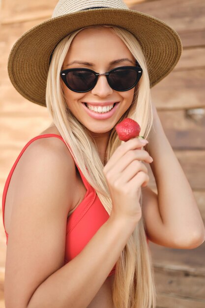 Młoda ładna uśmiechnięta blond kobieta w stroju kąpielowym, w okularach przeciwsłonecznych i kapeluszu, podczas gdy radośnie trzyma truskawkę na plaży