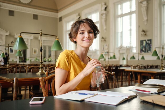 Młoda ładna uśmiechnięta studentka z wodą w ręku radośnie patrząca w kamerę podczas nauki w bibliotece uniwersyteckiej