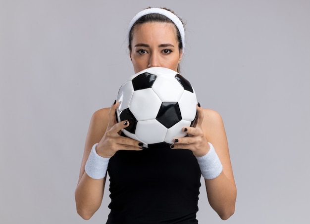 Młoda ładna wysportowana dziewczyna nosząca opaskę na głowę i opaski trzymająca piłkę nożną od tyłu