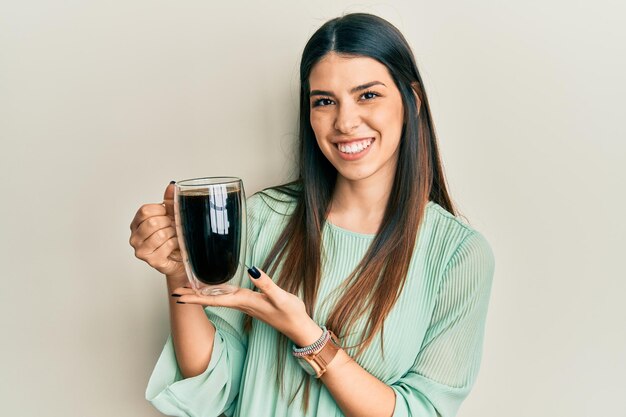 Młoda latynoska kobieta pije filiżankę kawy, wyglądając pozytywnie i szczęśliwie, stojąc i uśmiechając się z pewnym siebie uśmiechem pokazującym zęby