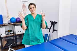 Bezpłatne zdjęcie młoda latynoska kobieta ubrana w mundur fizjoterapeuty stojąca w klinice, pokazująca i wskazująca palcami numer osiem, uśmiechając się pewnie i szczęśliwie.