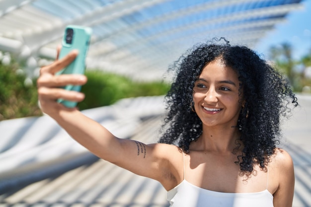 Młoda latynoska kobieta uśmiecha się pewnie robiąc selfie przy smartfonie w parku