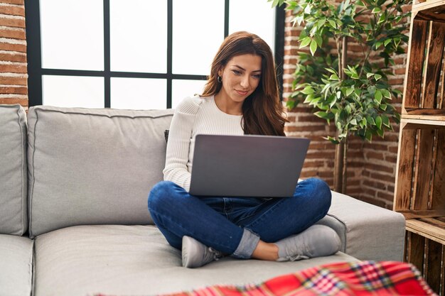 Młoda latynoska kobieta używa laptopa siedzi na kanapie w domu