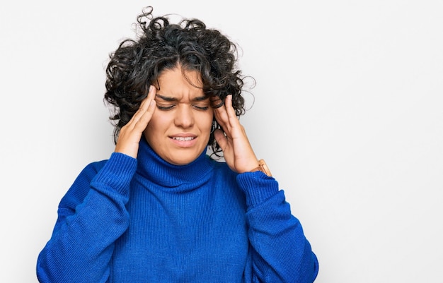 Młoda latynoska kobieta z kręconymi włosami ubrana w sweter z golfem z ręką na głowie z powodu stresu cierpiącego na migrenę