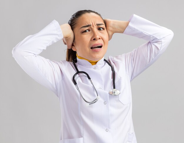 Młoda lekarka w białym fartuchu medycznym ze stetoskopem na szyi z poirytowanym wyrazem twarzy dotykającym jej głowy rękami