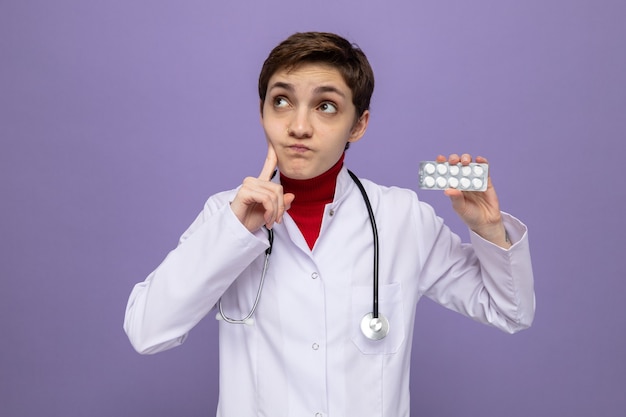 Młoda lekarka w białym fartuchu ze stetoskopem na szyi trzymająca blister z tabletkami patrząca w górę zdziwiona stojąc nad fioletową ścianą