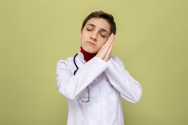 Młoda lekarka w białym fartuchu ze stetoskopem na szyi trzymająca się razem za ręce wykonująca gest snu opierając głowę na dłoniach z zamkniętymi oczami, stojąc na zielono