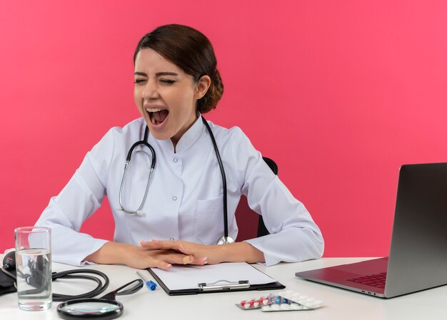 Młoda lekarka w szlafroku medycznym i stetoskopie siedzi przy biurku z narzędziami medycznymi i laptopem, kładąc ręce na biurku i krzycząc z zamkniętymi oczami na różowej ścianie