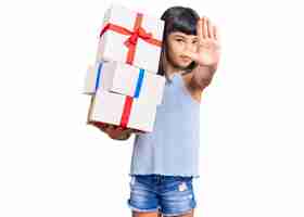 Bezpłatne zdjęcie młoda mała dziewczynka z hukiem trzyma prezenty z otwartą dłonią robi znak stop z poważnym i pewnym siebie gestem obrony