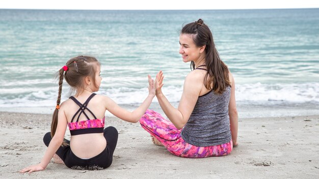 Młoda mama z małą córeczką w sportowej odzieży siedzi na plaży na tle morza. Wartości rodzinne i zdrowy tryb życia.