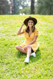 Młoda meksykańska kobieta w żółtej sukience i sombrero siedzi na trawie w parku.