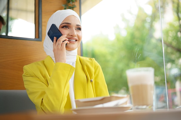 Młoda muzułmańska bizneswoman w chuście na głowie siedzi w kawiarni i rozmawia przez telefon