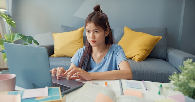 Młoda nastolatka z Azji z laptopem do codziennego użytku uczy się online pisać zeszyt wykładowy do testu końcowego w salonie w domu