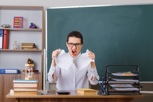 Młoda nauczycielka w okularach rozrywa kawałek papieru krzycząc z agresywnym wyrazem szalony i szalony siedzący w szkolnej ławce przed tablicą w klasie