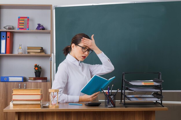 Młoda nauczycielka w okularach siedzi przy ławce szkolnej przed tablicą w klasie, czytając książkę, wyglądając na zdezorientowaną i zakłopotaną