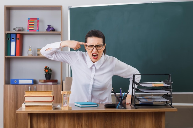 Bezpłatne zdjęcie młoda nauczycielka w okularach siedzi przy ławce szkolnej przed tablicą w klasie, patrząc na kamerę z agresywnym wyrazem twarzy