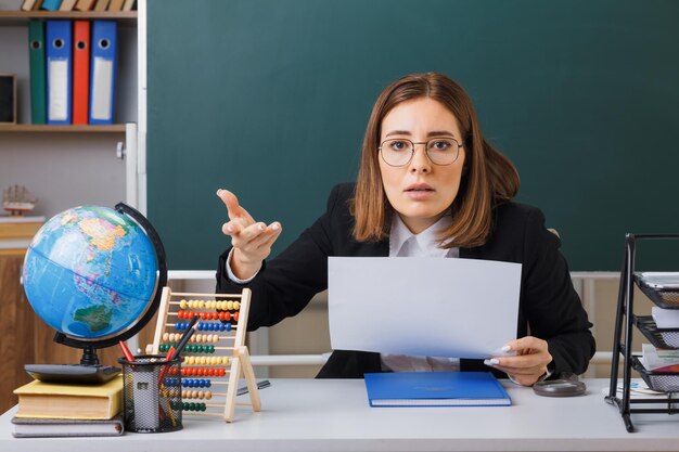 Młoda nauczycielka w okularach siedzi przy ławce szkolnej z kulą ziemską i książkami przed tablicą w klasie, trzymając białą pustą kartkę papieru podnoszącą rękę z niezadowoleniem i oburzeniem