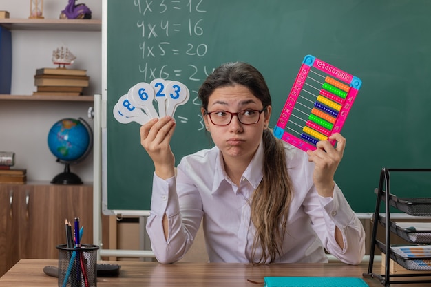 Młoda nauczycielka w okularach siedzi przy szkolnej ławce z rachunkami i tablicami rejestracyjnymi, wyglądająca na zmęczoną i znudzoną, dmuchająca w policzki, wyjaśniająca lekcję przed tablicą w klasie