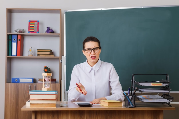 Młoda nauczycielka w okularach sprawdzająca pracę domową niezadowolonych uczniów siedzących w szkolnej ławce przed tablicą w klasie
