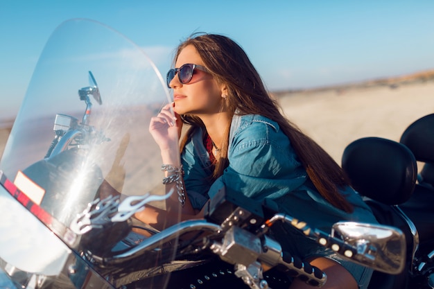 Młoda niesamowita seksowna kobieta siedząca na motocyklu na plaży, ubrana w stylowy crop top, koszule, doskonale dopasowana do szczupłego, oswojonego ciała i długich włosów. Portret na zewnątrz stylu życia.
