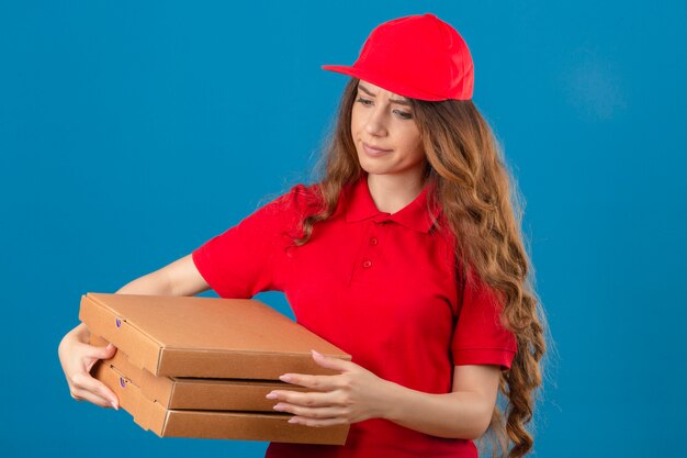 Młoda niezadowolona kobieta dostawy z kręconymi włosami, ubrana w czerwoną koszulkę polo i czapkę ze stosem pudełek po pizzy, patrząc na kamerę z nieszczęśliwą twarzą na izolowanym niebieskim tle