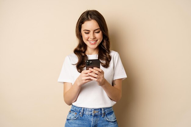 Młoda, nowoczesna dziewczyna rozmawiająca w aplikacji za pomocą aplikacji na smartfona i uśmiechająca się, stojąc na beżowym tle
