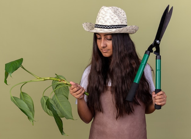 Młoda ogrodniczka dziewczyna w fartuch i letni kapelusz trzymając maszynkę do strzyżenia roślin i żywopłotu, patrząc pewnie stojąc nad zieloną ścianą