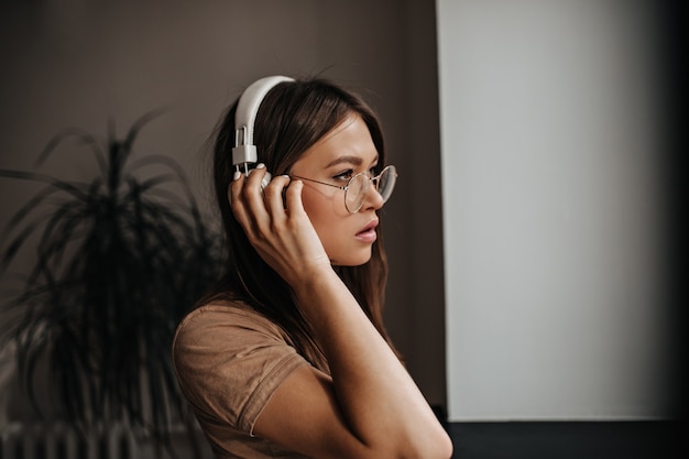 Młoda opalona kobieta w białych słuchawkach i okularach z poważnym wyglądem pozuje w pokoju.