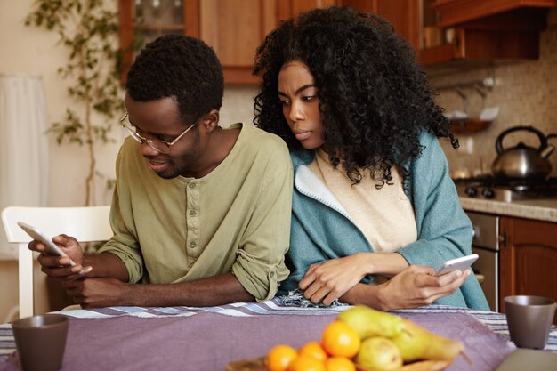 Młoda para Afroamerykanów korzystająca z elektronicznych gadżetów w domu: szczęśliwy mąż przeglądający wiadomości w mediach społecznościowych, podczas gdy jego zazdrosna zaborcza żona szpieguje, czyje zdjęcia lubi