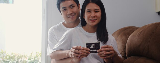 Młoda para azjatyckich w ciąży pokazać i patrząc zdjęcie USG dziecka w brzuchu. Mama i tata czują się szczęśliwi, uśmiechając się spokojnie, podczas gdy dbają o dziecko leżące na kanapie w salonie w domu.