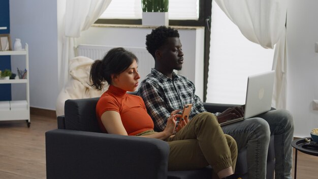 Młoda para etniczna multi za pomocą technologii w salonie w domu. Międzyrasowe osoby ze smartfonem i laptopem siedząc razem na kanapie. Żona i mąż rasy mieszanej z urządzeniami