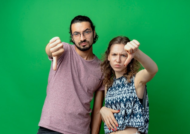 młoda para, mężczyzna i kobieta, niezadowolony, pokazując kciuki w dół stojąc nad zieloną ścianą