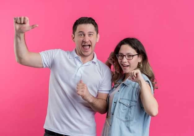 Młoda para mężczyzna i kobieta w ubranie szczęśliwy i podekscytowany, wskazując palcami na siebie, stojąc na różowej ścianie