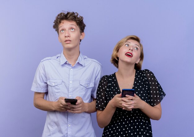 Młoda para piękny stojący obok siebie, trzymając smartfony, patrząc zdezorientowany na niebieskiej ścianie