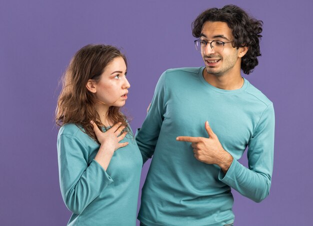 Młoda para pod wrażeniem kobiety uśmiechnięty mężczyzna ubrany w piżamę mężczyzna w okularach wskazujący na kobietę, która trzyma rękę na klatce piersiowej, patrząc na siebie na białym tle na fioletowej ścianie