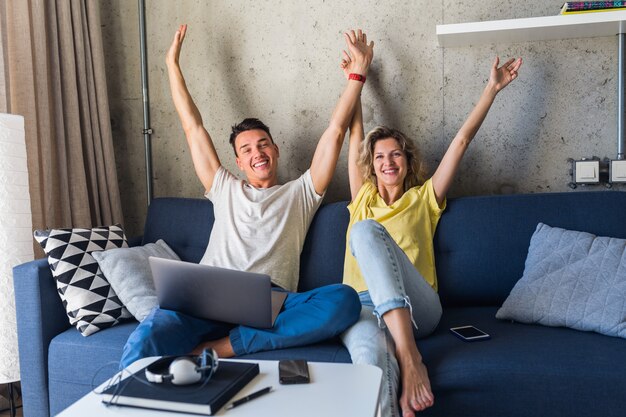 Młoda para siedzi na kanapie w domu, śmiejąc się, trzymając się za ręce, używając laptopa w Internecie
