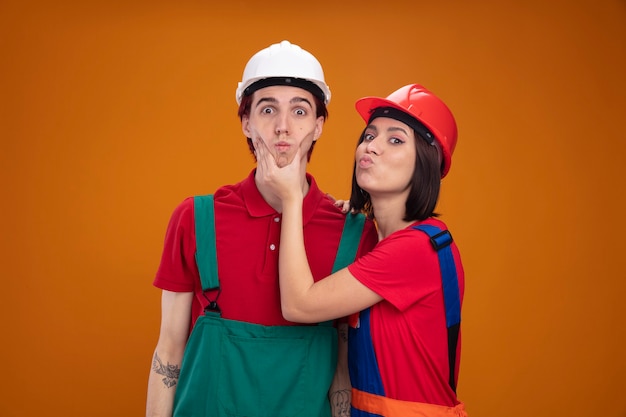 Młoda para w mundurze pracownika budowlanego i kasku dziewczyna stojąca w widoku profilu, chwytając policzki nieświadomego faceta, ściągając usta oba