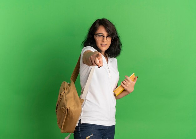Młoda pewna siebie całkiem kaukaska uczennica w okularach iz powrotem wskazuje na aparat trzymając notebook na zielono z miejsca na kopię