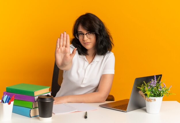 Młoda, pewna siebie, ładna kaukaska uczennica w okularach siedzi przy biurku z gestami narzędzi szkolnych stop znak ręką na pomarańczowo z miejscem na kopię