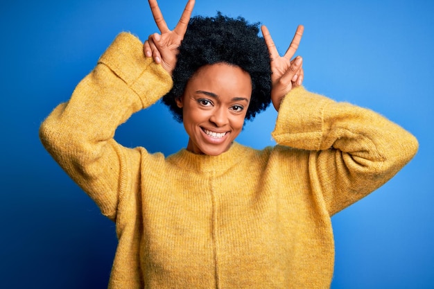 Bezpłatne zdjęcie młoda piękna afroamerykanka afro z kręconymi włosami, ubrana w żółty swobodny sweter, pozująca zabawnie i szalona z palcami na głowie jako królicze uszy uśmiechająca się wesoło